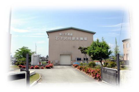 橙色の外壁に「国土交通省 石子沢川排水機場」と文字が貼り付けられている三角屋根で建物までの道には緑の植木やピンク色の花が咲いた花壇がある石子沢川排水機場の外観写真