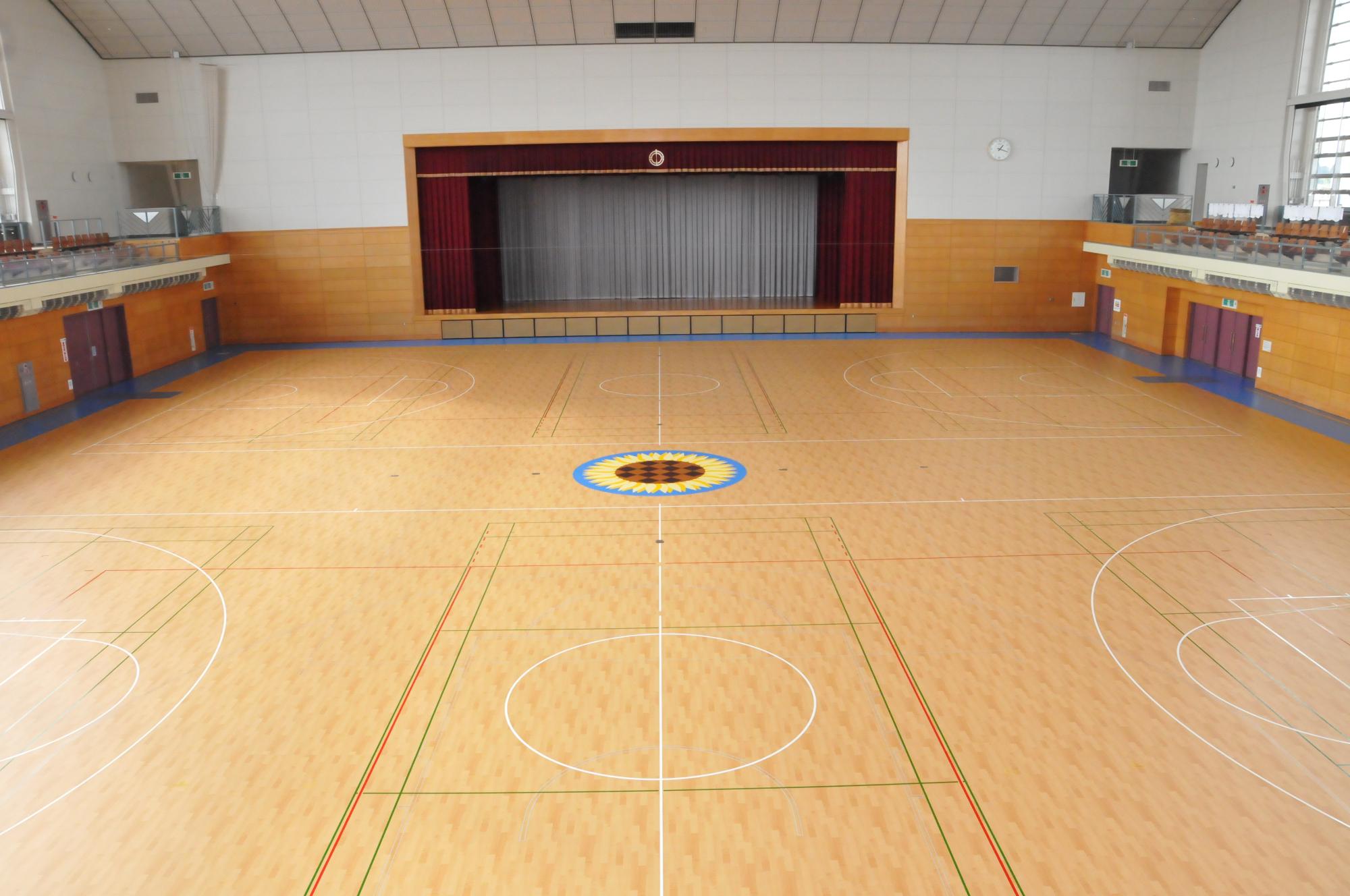 中央床にバスケットボールのコートが2面設置され、奥には深く赤い色の一文字幕や袖幕グレーの引幕の閉じたステージがあり、両側2階席に観客席が設けられている中山町体育館館内の写真
