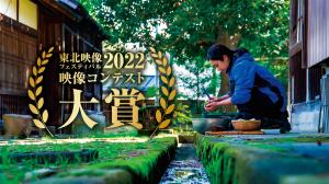 両側を月桂樹で囲み金色で書かれた「東北映像フェスティバル2022映像コンテスト大賞」の文字と青い服を着た女性が、緑溢れる小さな小川が流れる傍で右手に包丁を持ちながら左手にもった里芋の皮をむく様子の写真