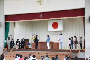 体育館の壇上で前方左側は椅子に座っている3名の先生方、前方中央で向かい合っている先生1名、手紙を読んでいる児童と花束を持っている児童の2名、そして前方右側に待機している5名の児童が写る離任式の様子の写真