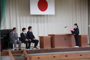 体育館壇上の前方左側で椅子に座っている3名の先生方に向かって、壇上の演台に置いてある立ちマイクの前で手紙を読んでいる児童の写真