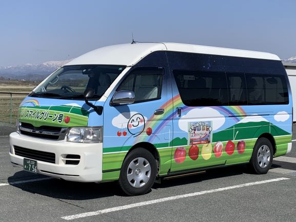 白を基調とした車体に、青空に虹がかかった山やりんごとさくらんぼのイラスト、「スマイルグリーン号」と正面に書かれている横向きで停車されたスマイルグリーン号の写真