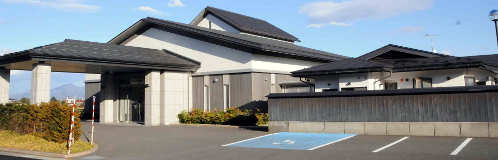 白い壁に濃いグレーの屋根の建物の玄関前に、屋根付きのロータリーがあり、右手に駐車スペースのある中山町斎場の外観写真