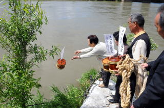 酒の入った小さな樽に白いのぼり旗を差し、水面から少し高さのある土手から投げ入れている男性と、その後ろで樽を持ち順番を待っている人達の写真