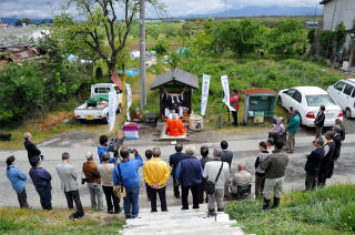 道路沿いにある金毘羅堂で神事が行われており、その様子を道の反対側に集まった人々が見守っている写真