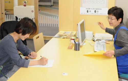 机の上で、右奥にデスクトップパソコンを前に青いエプロンを着用した女性職員が左手前にいる申込用紙を記入している女性に対して、口頭で説明している様子の写真