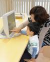 机の上に設置された備え付けのパソコンモニター画面を、男の子とお母さんが一緒に人差し指を指しながら本を探している様子の写真