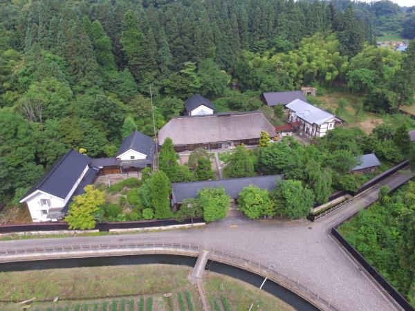 手前に道路、後ろに森があり、敷地内には大きな母屋や複数の蔵が立ち並び、たくさんの木が植えられた旧柏倉家住宅の全景写真