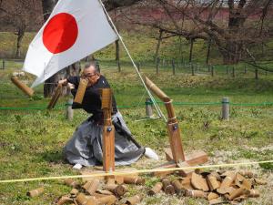 日の丸国旗が掲揚されたお達磨の桜公園敷地内で、黒袴姿の男性が真剣を使い、目の前に設置された2本の巻きわらを切った瞬間の写真