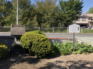 県道178号線を背景に、白い案内看板の周辺に、低木や草花が植えられ、三角屋根の小さな祠が佇んでいる写真