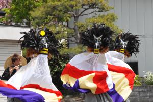 松の木が植えられた民家の前で、横笛を奏でる人の前で黒い獅子のお面を被った3名の踊り手が舞を披露している様子の写真