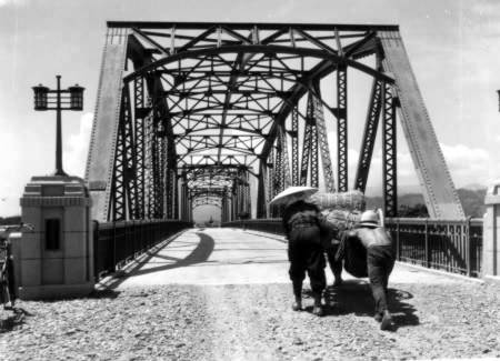 鉄筋コンクリートに作り替えられた長崎橋を、2人掛かりで大きな荷車を押しながら渡ろうとしている様子のモノクロ写真