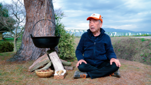 黒い鍋が吊るされた鍋掛松の根本に、竹ざるにはいった里芋と、四角い箱にはいった棒だらが置いてあり、その横には白とオレンジ色のキャップを被り青い服を着た年配の男性が座っている様子の写真