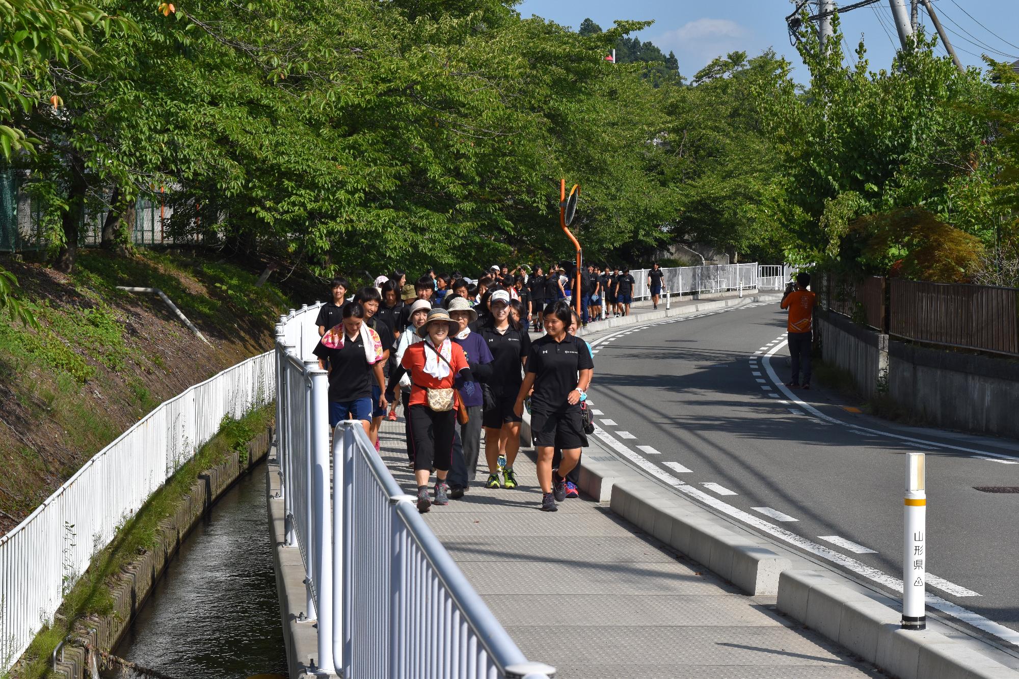 用水路沿いの歩道を、帽子やタオルを身に付けた大勢の参加者達が、列をなしてウォーキングしている様子の写真(なかやま健幸クラブ)