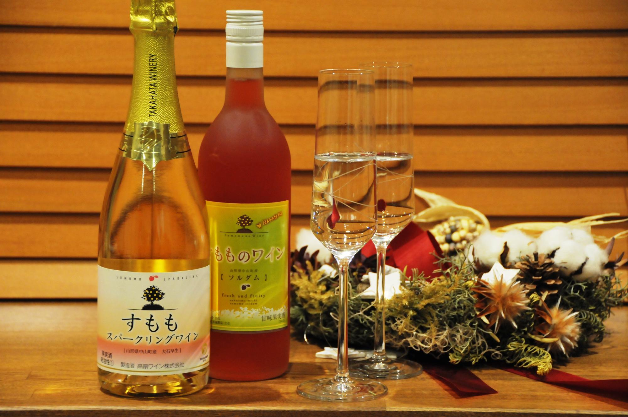 段差のついた木製の壁を背景に、木製のテーブルの上には左からすもものスパークワイン、すもものワイン（ソルダム）、中身の入った2本のシャンパングラス、飾られたクリスマスリースの順に飾られている様子の写真