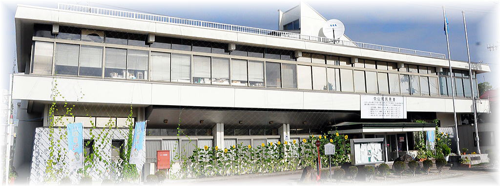 役場玄関横には掲示板が設置され、花壇にはヒマワリの花が並んで植えられており、二階のベランダに向け張られたネットに沿うようにつるを伸ばした緑のカーテンがある2階建ての中山町役場の外観写真